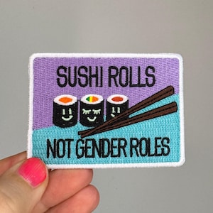 Könnte beinhalten: Ein rechteckiger bestickter Aufnäher mit violettem Oberteil und hellblauem Unterteil. Er zeigt drei Sushi-Rollen mit Gesichtern, zwei braune Essstäbchen und den schwarzen Text "SUSHI ROLLS NOT GENDER ROLES" auf der Oberfläche.