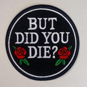 May include: Circular black patch with the text "BUT DID YOU DIE?" in white. Two red roses with green leaves are on either side of the text. The patch has a white border.