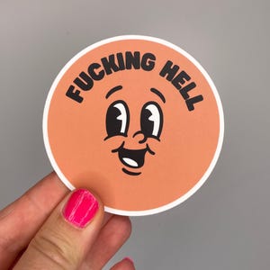 Könnte beinhalten: Ein runder Aufkleber mit einem orangefarbenen Cartoon-Gesicht und dem Text "FUCKING HELL" in Schwarz. Das Gesicht hat große schwarze Augen, einen lächelnden Mund und einen weißen Rand.