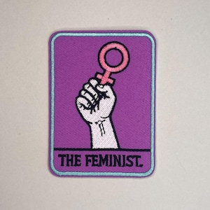 Peut inclure: Un écusson rectangulaire avec un fond violet et une bordure bleu clair. L'écusson représente un poing blanc tenant un symbole de Vénus rose. Le texte "THE FEMINIST" est affiché en blanc en bas.