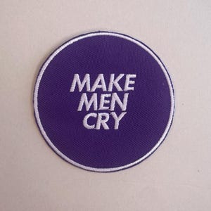 May include: A circular purple patch with the white embroidered text "MAKE MEN CRY." The patch has a white border. It is designed to be sewn or ironed onto clothing or accessories. The design is simple and bold, suitable for customisation.