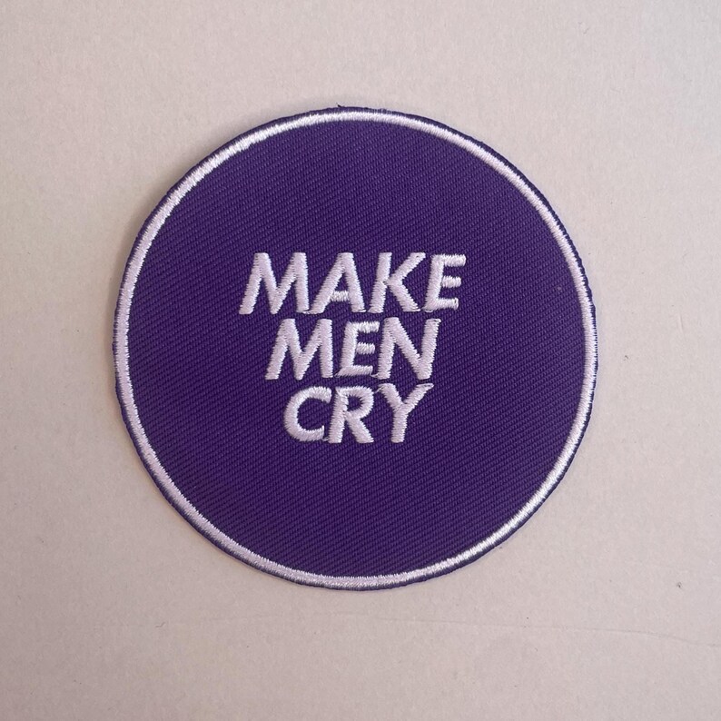 May include: A circular purple patch with the white embroidered text "MAKE MEN CRY." The patch has a white border. It is designed to be sewn or ironed onto clothing or accessories. The design is simple and bold, suitable for customisation.