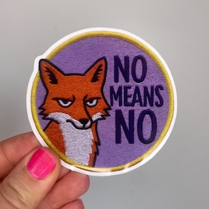 Könnte beinhalten: Runder Aufnäher mit einem orangefarbenen Fuchs mit ernstem Blick in einem lila Kreis. Die Worte "NO MEANS NO" sind in Dunkelblau gedruckt. Der Aufnäher hat einen gelben Rand und wird von einer Hand gehalten.