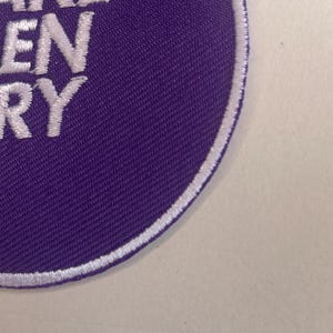 May include: A circular purple patch with white embroidered text that reads "TAKE GREEN VERY". The patch has a white border and is set against a light background. The patch is likely for clothing or accessories.