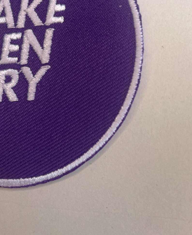May include: A circular purple patch with white embroidered text that reads "TAKE GREEN VERY". The patch has a white border and is set against a light background. The patch is likely for clothing or accessories.