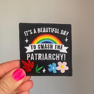 Puede incluir: Parche cuadrado negro con el texto "IT'S A BEAUTIFUL DAY TO SMASH THE PATRIARCHY!" sobre un arcoíris. El parche tiene flores y hojas bordadas en rojo, azul y rosa.