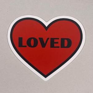 Könnte beinhalten: Ein herzförmiger Aufkleber mit roter Mitte und dem Wort "LOVED" in schwarzen Blockbuchstaben. Das Herz hat einen schwarzen Umriss und einen weißen Rand. Der Aufkleber befindet sich auf einem hellgrauen Hintergrund.