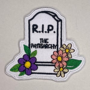Könnte beinhalten: Gestickter Aufnäher mit einem Grabstein-Design und dem Text "R.I.P. THE PATRIARCHY". Der Aufnäher ist weiß mit schwarzer Schrift und einem schwarzen Rand. Bunte Blumen in Rosa, Gelb und Lila mit grünen Blättern schmücken den unteren Teil des Grabsteins.