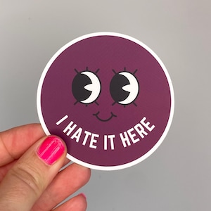 Könnte beinhalten: Ein runder Aufkleber mit lila Hintergrund und weißem Rand. Der Aufkleber zeigt ein Cartoon-Gesicht mit großen weißen Augen, schwarzen Pupillen und einem kleinen Lächeln. Der Text "I HATE IT HERE" ist weiß gedruckt.