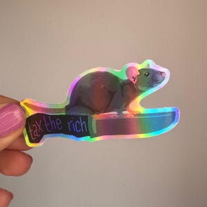 Könnte beinhalten: Ein holografischer Aufkleber mit einer dunkelgrauen Ratte, die auf einem großen Messer sitzt. Die Messerklinge ist schwarz und trägt den Text "tax the rich" in violetten Buchstaben. Der gesamte Aufkleber hat einen reflektierenden Regenbogen-Glanz.