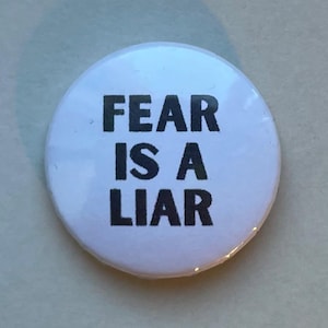 Può includere: Un bottone bianco rotondo con la scritta "FEAR IS A LIAR" in grassetto e maiuscolo nero. Il bottone ha una finitura lucida. Il testo è centrato sul bottone.