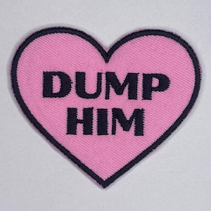 Könnte beinhalten: Ein pinkfarbener herzförmiger Aufnäher mit schwarzem, gesticktem Text „DUMP HIM“. Der Aufnäher hat einen schwarzen Rand und besteht aus einem strukturierten Stoff. Geeignet für Kleidung oder Accessoires.
