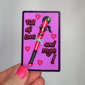 Könnte beinhalten: Ein rechteckiger bestickter Aufnäher mit violettem Hintergrund zeigt eine rote Baseballschläger-Figur mit Gesicht, schwarzen Haaren und einer hellblauen Fliege. Der Text Full of Love and rage! ist von kleinen roten Herzen umgeben.