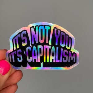 May include: Holographic sticker with the text "IT'S NOT YOU IT'S CAPITALISM" in a bold, multi-coloured font. The sticker has a black border and a rainbow sheen, held by a hand with pink nail polish.