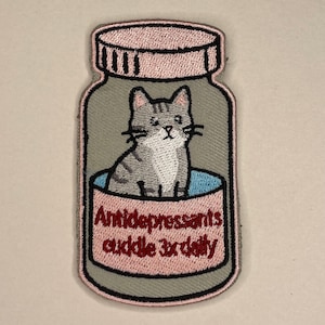 Peut inclure: Écusson brodé en forme de flacon de pilules, représentant un chat tigré gris à l'intérieur. L'écusson a des accents roses et gris, avec les mots "Antidépresseurs câlins 3x par jour" en texte rose.