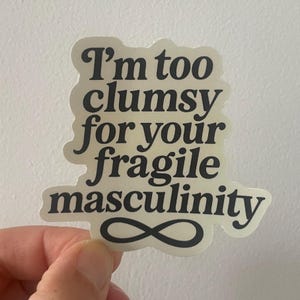 Könnte beinhalten: Cremefarbener Aufkleber mit dem Text "I'm too clumsy for your fragile masculinity" in fetter, schwarzer Schrift. Das Design enthält ein schwarzes Unendlichkeitszeichen.