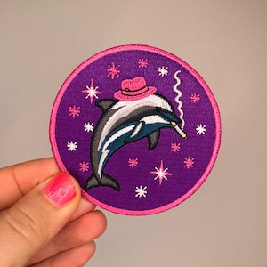 Könnte beinhalten: Ein runder bestickter Aufnäher mit einem grau-weißen Delfin, der einen pinken Fedora-Hut trägt. Der Delfin hält eine brennende Zigarette im Mund, aus der Rauch aufsteigt. Der Hintergrund ist violett und mit pinken und weißen Sternen verziert.