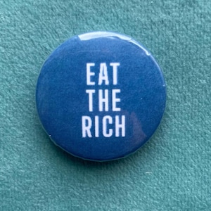 Può includere: Un bottone rotondo blu con il testo bianco "EAT THE RICH" in grassetto, senza grazie. Il bottone ha una finitura lucida ed è posto su uno sfondo strutturato, verde acqua.