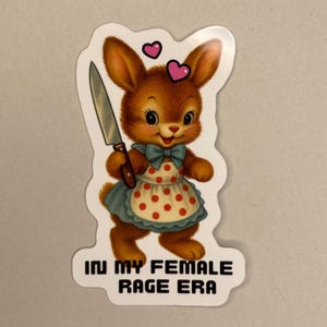 Könnte beinhalten: Ein Aufkleber mit einer Retro-Illustration eines braunen Hasen, der ein Messer hält und ein blaues Kleid und eine rot gepunktete Schürze trägt. Zwei rosa Herzen schweben über dem Hasen. Der Text "IN MY FEMALE RAGE ERA" steht unten.
