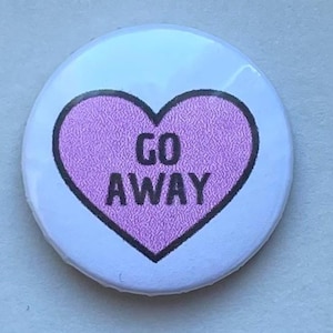 Può includere: Un bottone rotondo con sfondo azzurro chiaro e un cuore rosa strutturato al centro. Il cuore contiene le parole "GO AWAY" in nero, con carattere sans-serif.