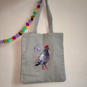 Könnte beinhalten: Eine hellgraue Tragetasche mit einer Grafik einer Taube, die ein rotes Barett und rosa Cowboystiefel trägt. Neben der Taube befinden sich eine Discokugel und der Text "IF I'M TOO MUCH... GO FIND LESS!". Die Tasche hat zwei Schulterriemen.