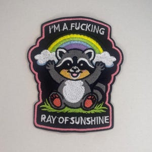 Könnte beinhalten: Gestickter Aufnäher mit einem Cartoon-Waschbären, Regenbogen und Wolken. Der Aufnäher trägt die Aufschrift "I'M A FUCKING RAY OF SUNSHINE" in Rosa und Schwarz. Der Waschbär ist grau, weiß und schwarz, mit rosa Akzenten.
