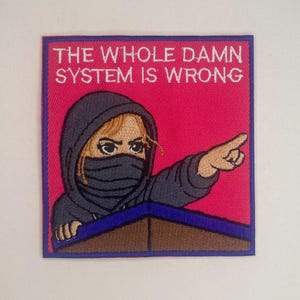 Könnte beinhalten: Gestickter Aufnäher mit dem Text "THE WHOLE DAMN SYSTEM IS WRONG" auf pinkfarbenem Hintergrund. Der Aufnäher zeigt eine Person in einem grauen Kapuzenpullover und Gesichtsbedeckung, die nach rechts zeigt. Der Aufnäher hat einen lila Rand.