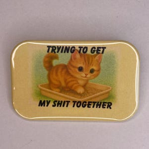 May include: A rectangular pin with a yellow background and a cartoon image of an orange tabby kitten in a litter box. The text reads "TRYING TO GET MY SHIT TOGETHER" in black letters.