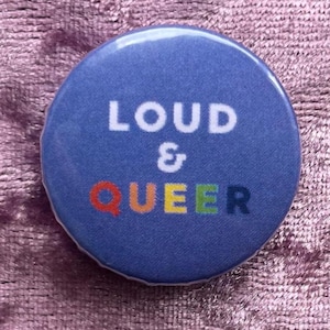Könnte beinhalten: Ein runder, blauer Button mit den Worten "LOUD & QUEER" in Weiß und Regenbogenfarben. Der Button ist ein Ansteckbutton, wahrscheinlich für eine Jacke, Tasche oder ein anderes Accessoire.