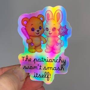 Op de afbeelding: Holografische sticker met een cartoonbeer en -konijn, elk gewapend. De beer houdt een roze knuppel vast, het konijn een knots. Tekst: "The patriarchy won't smash itself!" Een kleurrijk, opvallend ontwerp.