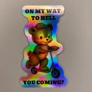 Può includere: Adesivo olografico con un orsetto dei cartoni animati su uno scooter. L'orso è marrone con un cappello verde e sorride. L'adesivo ha il testo "ON MY WAY TO HELL" in alto e "YOU COMING?" in basso.