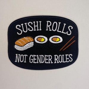 Könnte beinhalten: Schwarzer, gestickter Aufnäher mit dem Text "SUSHI ROLLS NOT GENDER ROLES". Der Aufnäher zeigt Illustrationen von Sushi-Rollen und Essstäbchen in Weiß, Orange und Braun.