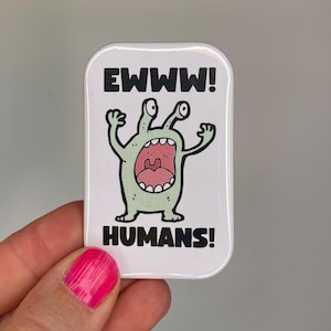 Könnte beinhalten: Ein weißer rechteckiger Button mit einer Cartoon-Alien-Illustration. Der Alien ist hellgrün mit großen Augen und offenem Mund. Der Text "EWww!" und "HUMANS!" sind in Schwarz.