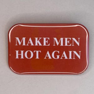 Può includere: Un bottone rettangolare rosso con angoli arrotondati. Il testo bianco "MAKE MEN HOT AGAIN" è centrato sul bottone. Il bottone ha una finitura lucida e un bordo bianco.