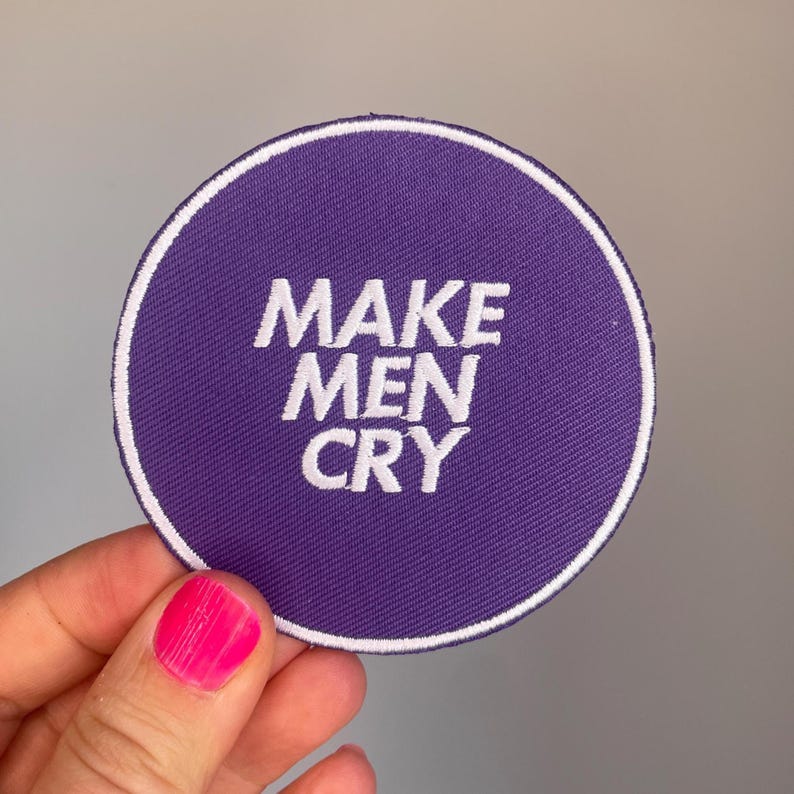 May include: A circular purple patch with the white embroidered text "MAKE MEN CRY." The patch has a white border. The patch is held by a hand with a pink painted fingernail.
