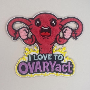 Peut inclure: Écusson brodé représentant un utérus de dessin animé avec les bras levés en position de flexion. L'utérus est rouge avec des contours noirs et une bordure blanche. Le texte "I LOVE TO OVARYact" est en dessous dans un design jaune et violet.