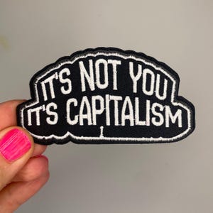 Könnte beinhalten: Schwarz-weißer, gestickter Aufnäher mit dem Text "IT'S NOT YOU IT'S CAPITALISM". Der Aufnäher hat einen schwarzen Hintergrund, weißen Text und einen weißen Rand. Der Aufnäher wird von einer Person mit pinkfarbenem Nagellack gehalten.
