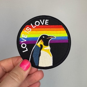 Könnte beinhalten: Ein runder, bestickter Aufnäher mit einem Pinguin vor schwarzem Hintergrund und einem horizontalen Regenbogenstreifen in der Mitte. Der weiße Schriftzug "LOVE IS LOVE" ist entlang des oberen linken Randes des schwarz umrandeten Aufnähers gebogen.