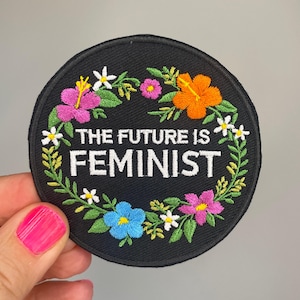 Può includere: Toppa rotonda ricamata nera con la scritta "THE FUTURE IS FEMINIST" circondata da una ghirlanda floreale. I fiori sono nei toni del rosa, arancione, blu e bianco.