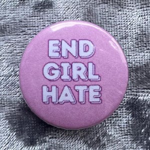 May include: A round, pink button with the words "END GIRL HATE" in white, outlined letters. The button is set against a textured, silver background. The button is a statement piece.