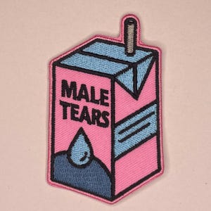 Könnte beinhalten: Rosa Aufnäher in Form eines Milchkartons mit den Worten "MALE TEARS" in schwarzen Blockbuchstaben. Der Karton hat blaue Akzente und eine blaue Tränen-Grafik.