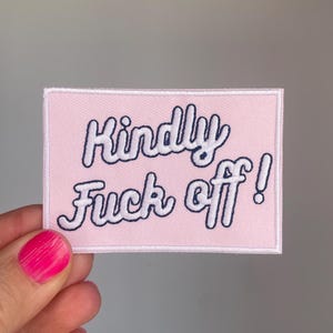 May include: A rectangular pink patch with the words "Kindly Fuck off!" embroidered in white script with a navy blue outline. The patch is held by a hand with a pink painted fingernail.