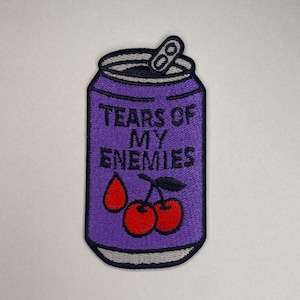 Könnte beinhalten: Gestickter Aufnäher in Form einer lila Getränkedose mit dem Text "TEARS OF MY ENEMIES" in Weiß. Die Dose zeigt zwei rote Kirschen und einen einzelnen roten Tropfen. Die Oberseite der Dose ist schwarz und silber.