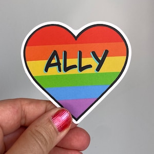 Könnte beinhalten: Ein herzförmiger Aufkleber mit horizontalen Streifen in Rot, Orange, Gelb, Grün, Blau und Violett. Das Wort ALLY ist in fetten schwarzen Großbuchstaben in der Mitte des Regenbogen-Designs aufgedruckt und mit einem dünnen weißen Rand versehen.