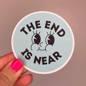 Könnte beinhalten: Ein runder Aufkleber mit hellblauem Hintergrund und dem Text "THE END IS NEAR" in Schwarz. Der Aufkleber zeigt ein Cartoon-Gesicht mit großen Augen und einem lächelnden Mund. Der Aufkleber wird von einer Hand mit pinkfarbenem Nagellack gehalten.