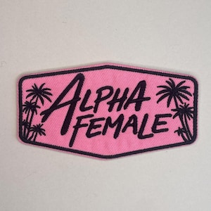 Może przedstawiać: Różowa naszywka z haftem z napisem "ALPHA FEMALE" w ciemnofioletowych literach. Naszywka ma czarną obwódkę i sylwetki palm po bokach.