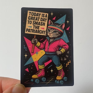 May include: A sticker showing a cat in athletic attire on roller skates, holding a sign saying "TODAY IS A GREAT DAY TO SMASH THE PATRIARCHY." The cat wears a pink jacket, blue trousers, and a rainbow headband, set against a black background with geometric shapes.