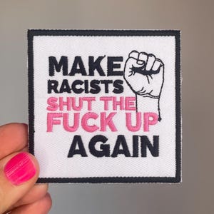 Könnte beinhalten: Quadratischer weißer Aufnäher mit schwarzem Rand. Der Aufnäher zeigt den Text "MAKE RACISTS SHUT THE FUCK UP AGAIN" in Schwarz und Pink, zusammen mit einer schwarzen Faust-Grafik. Der Aufnäher ist wahrscheinlich zum Aufbügeln oder Aufnähen.