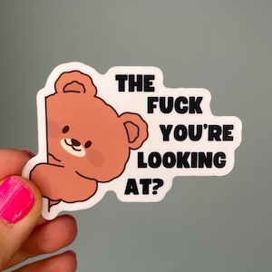 Könnte beinhalten: Ein Aufkleber mit einem Cartoon-Bären, der von der linken Seite hervorlugt. Der Bär ist hellbraun mit einem lächelnden Gesicht. Der Text in fetter schwarzer Schrift lautet "THE FUCK YOU'RE LOOKING AT?" auf weißem Hintergrund.
