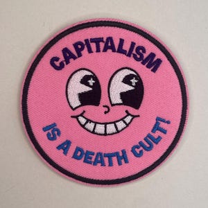 May include: A round, pink patch with the words "CAPITALISM IS A DEATH CULT!" in blue. The patch features a smiling face with white eyes and teeth, and a black outline.